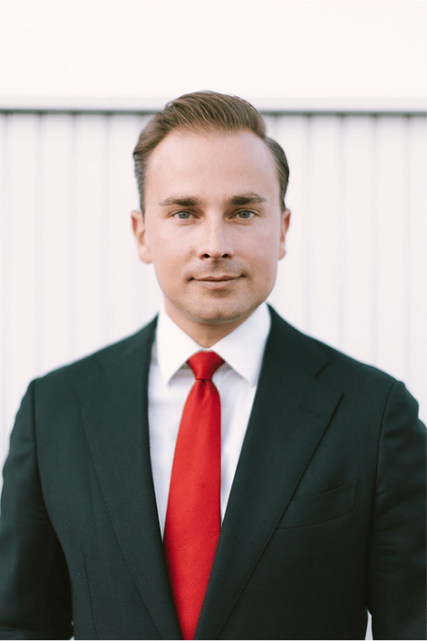 Professional headshot: man in a dark suit, white shirt, and red tie, facing the camera with a neutral expression against a light background.